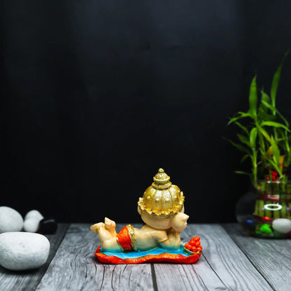 Aurance handcrafted resin Bal Ganesha figurine lying down and reading the Ramayana on a wooden surface with bamboo