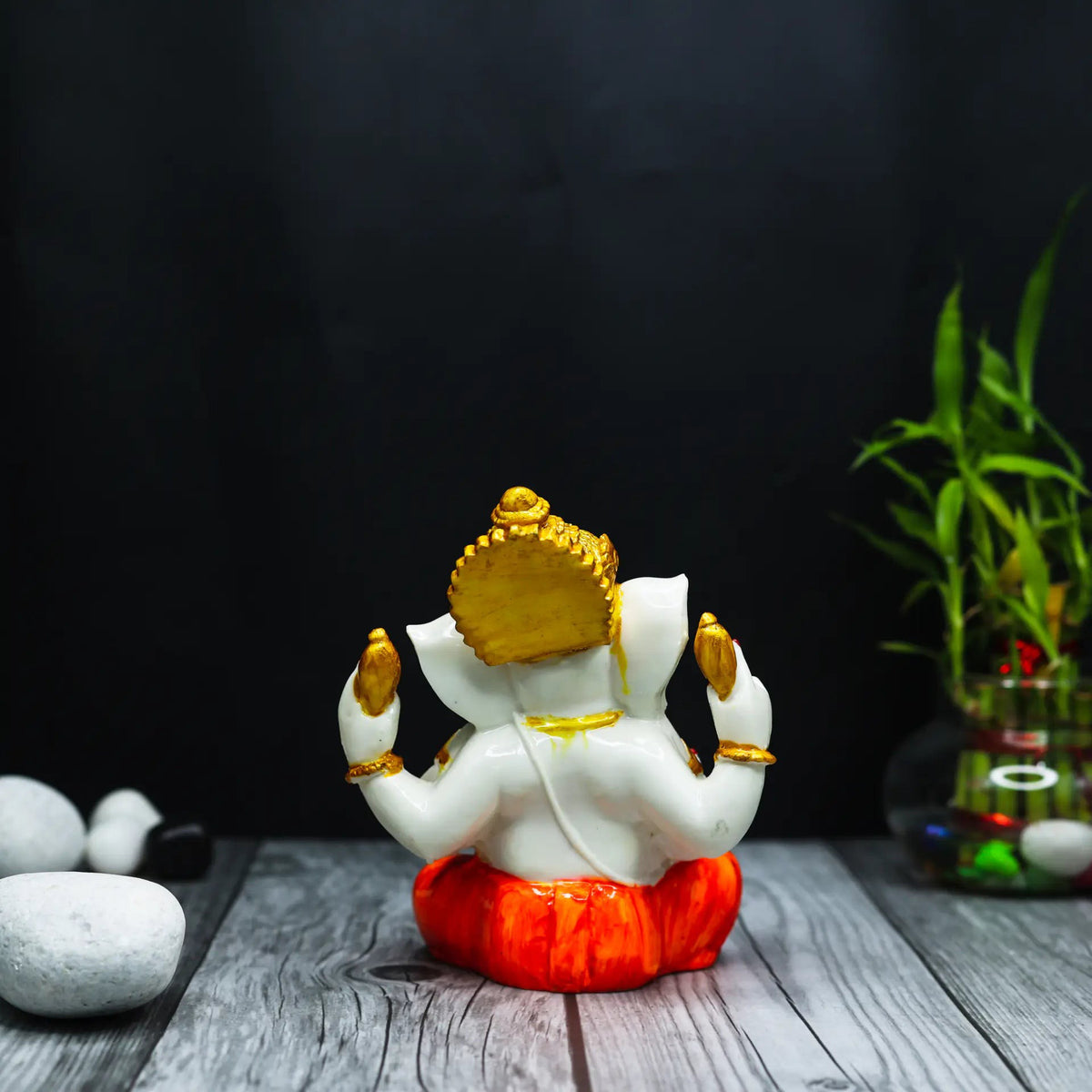 Rear view of an Aurance Ganesha idol with white body, gold crown, and orange base, set against a black background.